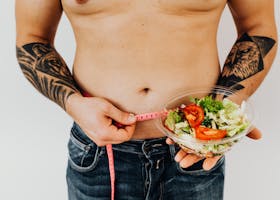 Tattooed man measuring waist while holding fresh salad, symbolizing healthy lifestyle and weight management.